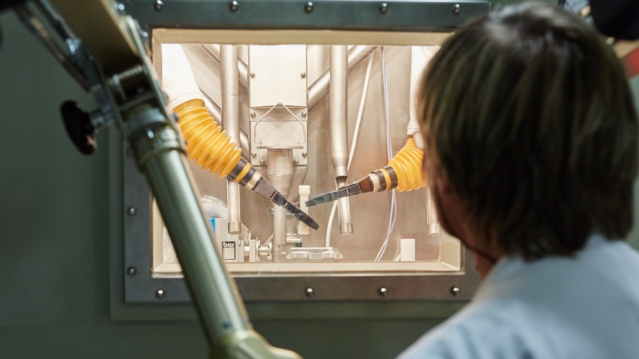 Ein Wissenschaftler schaut durch ein Fenster in eine verschlossene Kammer, in der zwei Roboterarme von der Decke hängen