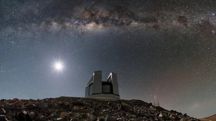 Auf einem steinigem Untergrund steht ein Teleskopgebäude. Es ist Nacht, im Hintergrund scheint der Vollmond und ein deutlich sichtbarer Sternenhimmel breitet sich über dem Bau aus.
