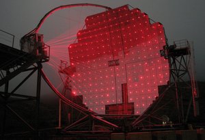 Ein parabolförmiger Teleskopspiegel mit roten Leuchtpunkten auf jedem Spiegelsegment. Zu den Leuchtpunkten führen rote Linien von einem Instrument links im Bild, das dem Spiegel gegenübersteht.