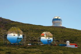 An einem Berghang stehen zwei große Teleskope mit Blick auf Parabolspiegel. Im Hintergrund ist eine weitere Teleskopkuppel zu sehen.
