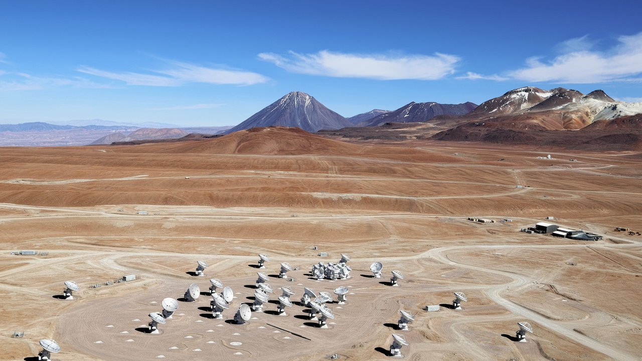 Luftaufnahme mehrerer weißer Antennenschüsseln in einer Wüstenlandschaft, im Hintergrund ein Gebirge