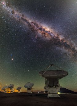 Sterne und Gasnebel in einem engen Band bilden die Milchstraße. Im Vordergrund stehen 6 der 66 ALMA-Teleskope, die zum Himmel gerichtet sind.