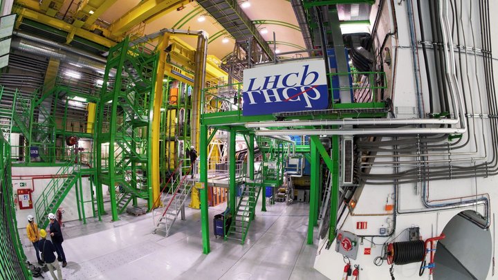 Große Halle mit Maschinen, die durch Leitungen und Rohre miteinander verbunden sind; im Vordergrund stehen drei Menschen mit Helmen