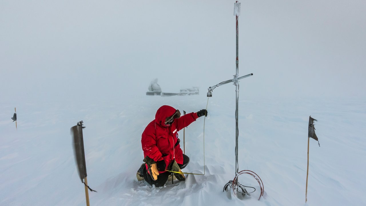 Wissenschaftler in rotem Anorak kniet im Schnee und hält eine antennenähnliche Konstruktion fest