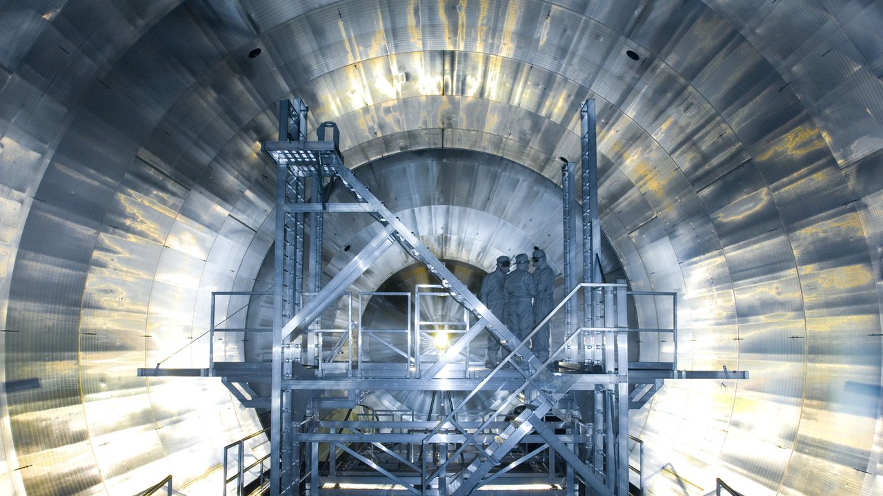 Im Inneren eines metallenen Tanks stehen drei Wissenschaftler in Schutzanzügen; eine Lampe in der Mitte erleuchtet die Szenerie
