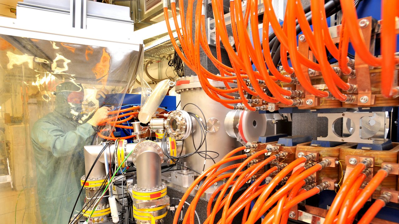 Rückwand eines Gerätes, in dem viele orangene kabel stecken; im Hintergrund ein Wissenschaftler im Schutzanzug