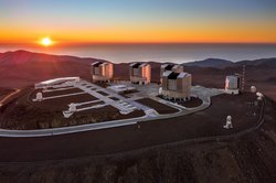 Die Gebäude des Teleskops auf einem Berg in der Wüste; im Hintergrund geht die Sonne auf