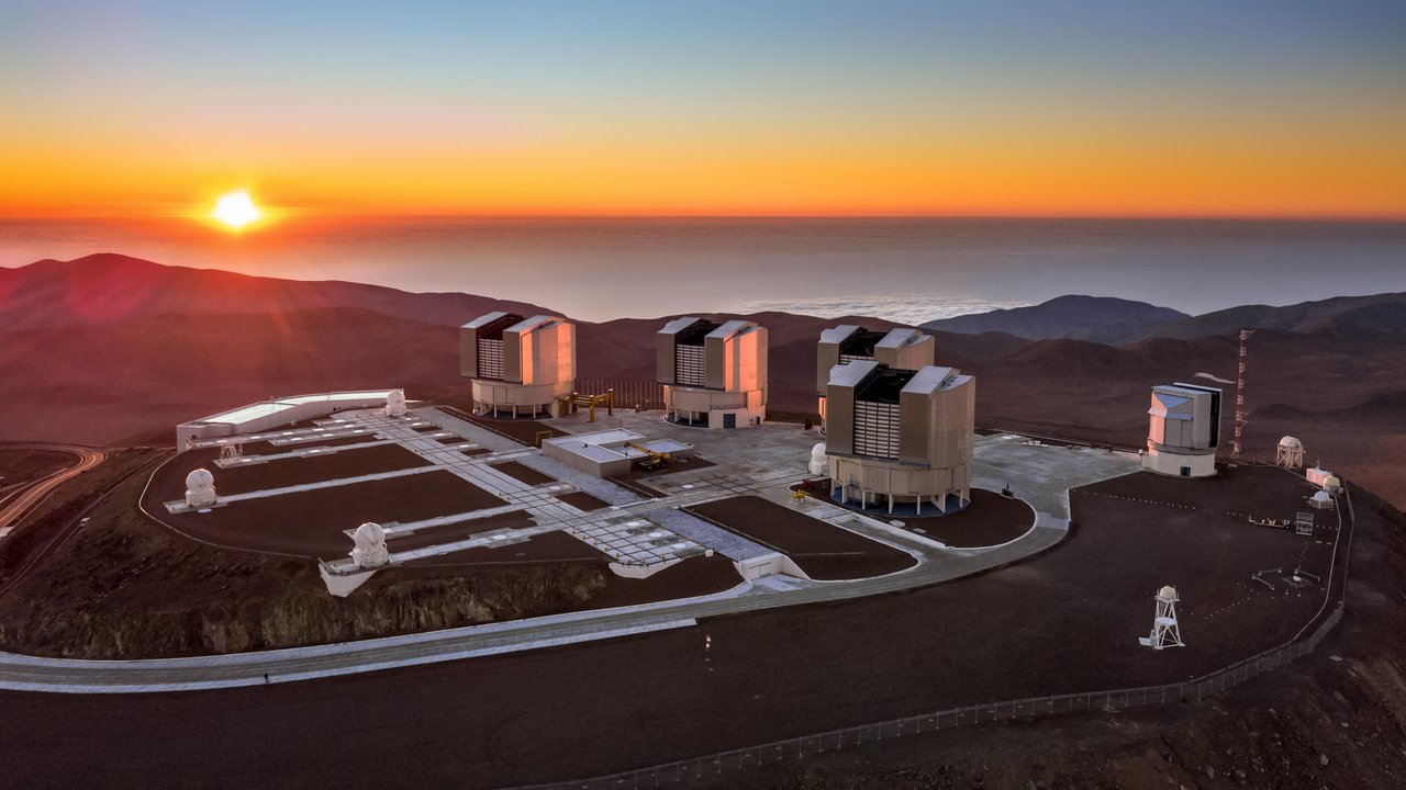 Die Gebäude des Teleskops auf einem Berg in der Wüste; im Hintergrund geht die Sonne auf