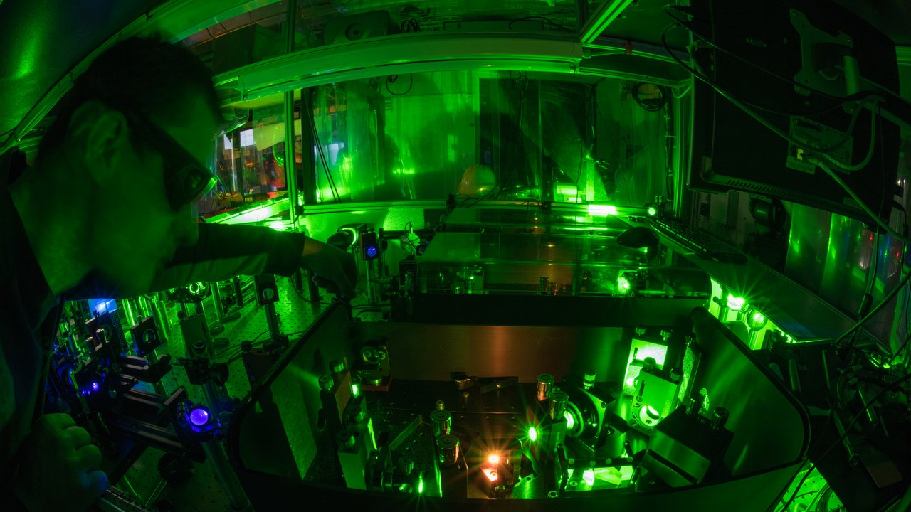 Wissenschaftler mit Schutzbrille beugt sich in einen Kasten, in dem mehrere Apparaturen festgeschraubt sind; es leuchten grüne und rote Lichter im abgedunkelten Raum