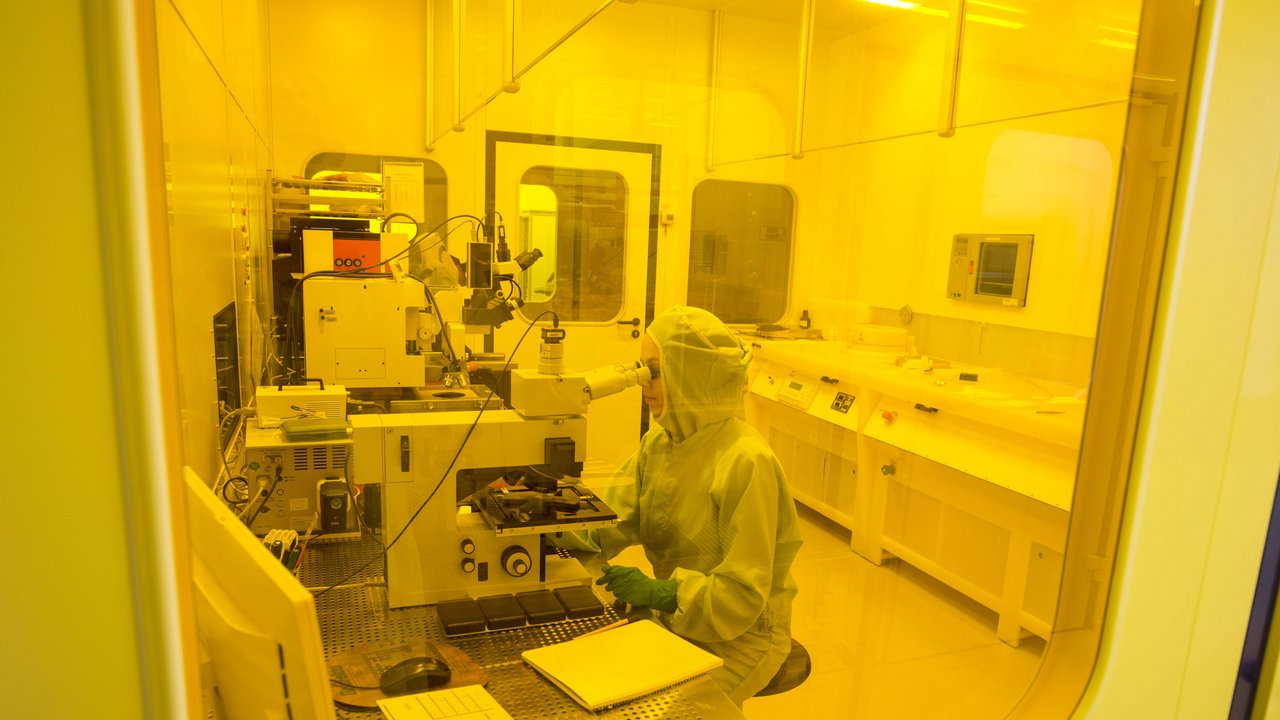 Eine Wissenschaftlerin sitzt in einem abgeschlossenen Raum vor einem Mikroskop und schaut hinein, die Szenerie ist in gelbes Licht getaucht, da sie durch eine gelbe Schutzfolie fotografiert wurde