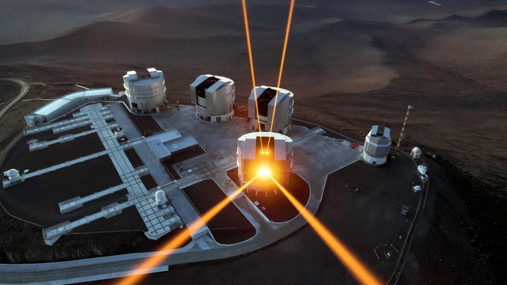 Teleskopanlage auf einem Berg in der Wüste; von einem Gebäude gehen vier Laserstrahlen in den Himmel