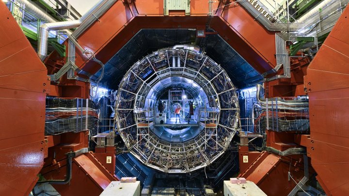 In einer mehreckigen, runden Öffnung befindet sich eine kreisrunde Röhre, die aus Metallgestänge besteht und im Inneren blau leuchtet; an ihrem anderen Ende steht ein Mensch.