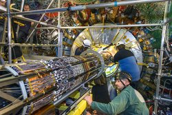 zwei Wissenschaftler arbeiten an einer metallenen, aus vielen einzelnen Verstrebungen bestehende Röhre, die in eine angeleuchtete Scheibe reicht