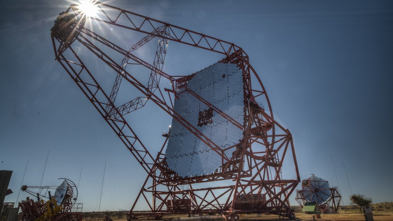 Ein großes Teleskop , in dem viele kleine Spiegelsegmente in einem Gerüst eingebaut sind im Vordergrund auf Wüstenboden und zwei kleinere ebensolche Teleskope im Hintergrund